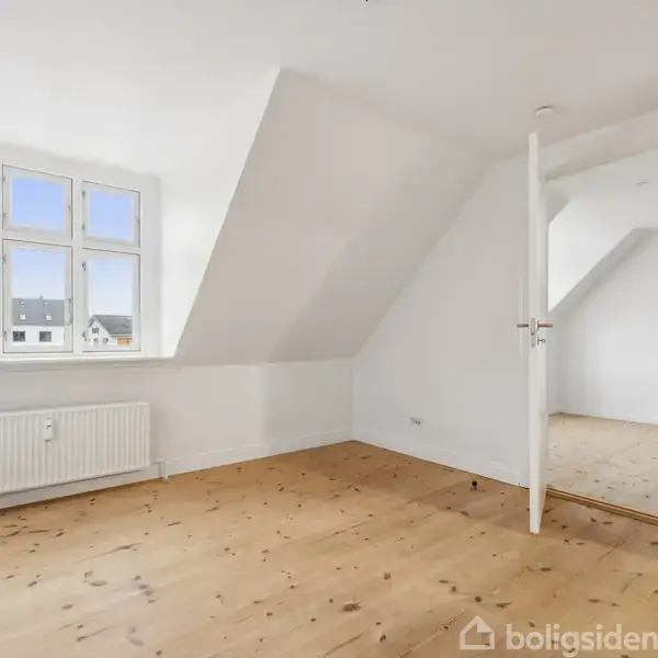 Et lyst værelse med trægulv, vindue med udsigt til nabobygninger, skråvægge og en åben dør til et andet rum. Radiator placeret under vinduet.