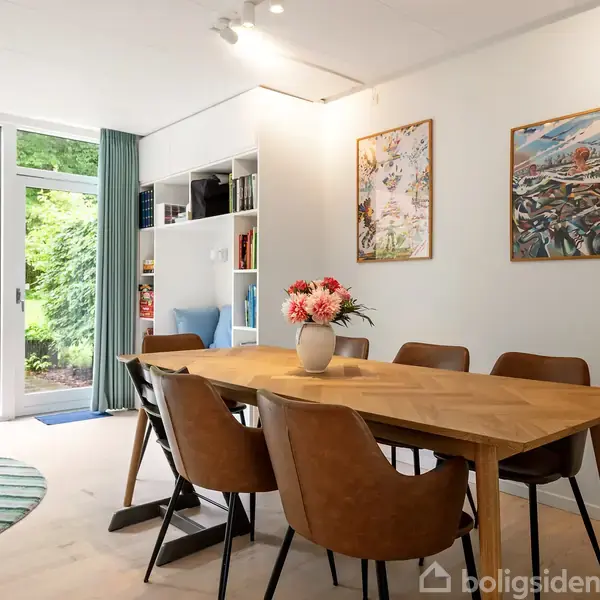 Spisebord med seks stole er centralt placeret, dekoreret med en vase blomster; værelse med lyse vægge, to kunstværker og en hylde. Store vinduer viser en grøn have.