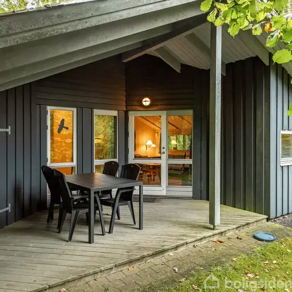 Sommerhus med skråtag vist udefra. En træveranda har et bord med fire sorte stole. Omgivet af grønne træer skaber en hyggelig atmosfære i skovområdet.