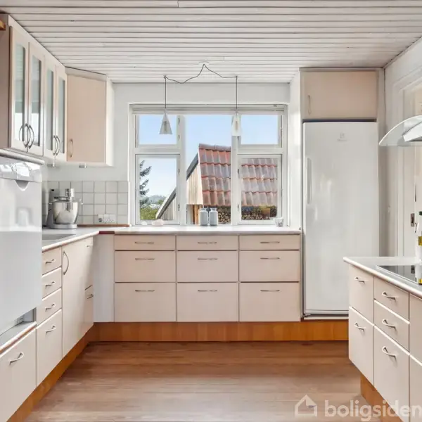 Køkken med hvide skabe og skuffer, rummeligt og moderne, placeret i et lyst rum med trægulv. Store vinduer åbner mod udendørs udsigt og tilføjer naturligt lys.