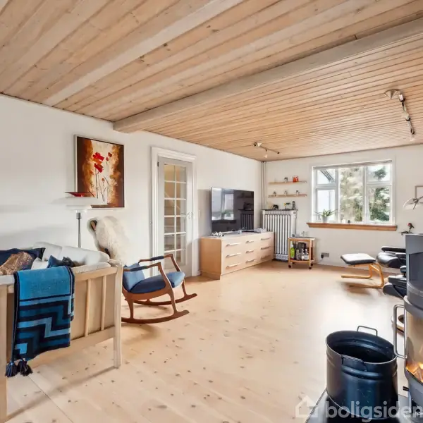 Sofa med puder og tæppe står ved siden af en gyngestol i en stue med lyst trægulv og loft. Pejs brænder til højre, og der er vinduer mod haven.