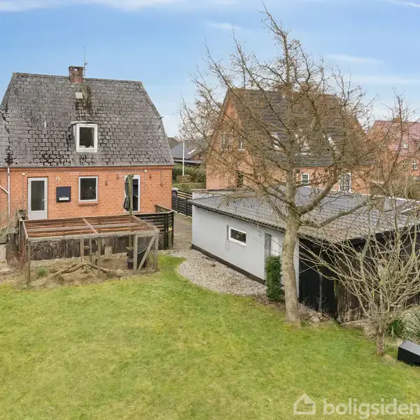 Rødt murstenshus med sort tag står i en have med græsplæne og hække. Ved siden af er et skur og et træ. Omgivet af andre lignende huse i et beboelsesområde.