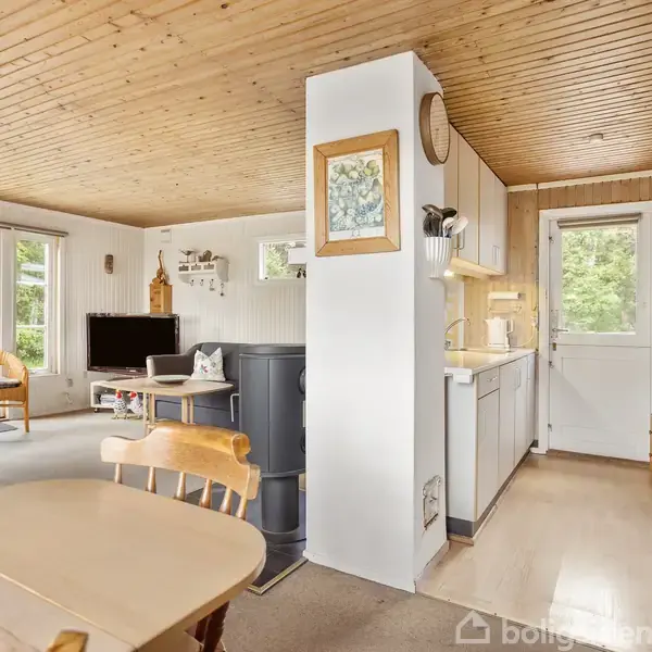 Stue med et træspisebord i forgrunden, en sort brændeovn i midten, og et TV-stueområde med sofagruppe i baggrunden. Store vinduer giver udsigt til grøn have.