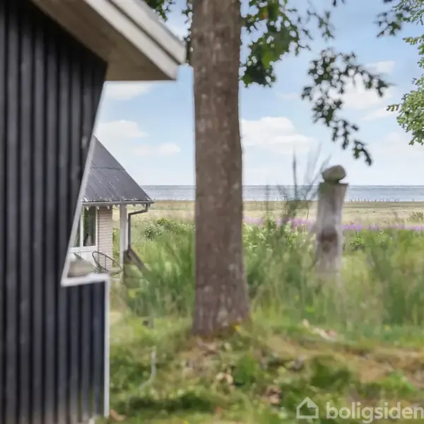 Et sort træhus står stille ved siden af en træstamme, mens grøn vegetation og en fjern havudsigt skaber en rolig, naturskøn baggrund.