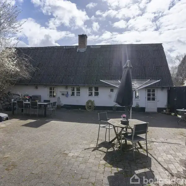 En gammel, hvidkalket bygning med stråtag står på en brolagt gårdsplads, hvor der er anrettet loungemøbler og parasoller. Der er træer i blomst i baggrunden under en skyet himmel.