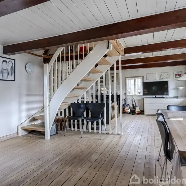 Trætrappe fører opad i en lys stue med trægulve, hvor der står et spisebord og stole. Vægge er dekoreret med billeder og en guitar står ved fjernsynet.