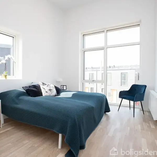 Soveværelse med en seng dækket af et blåt tæppe, stående ved siden af et lille bord med lampe. Et vindue og en dør giver udsigt til bygninger udenfor.