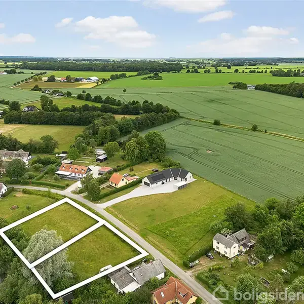 Grønne marker omkranser en række huse ved en sø. Et markeret jordstykke vises i forgrunden. Store åbne marker strækker sig mod horisonten under en blå himmel med lette skyer.