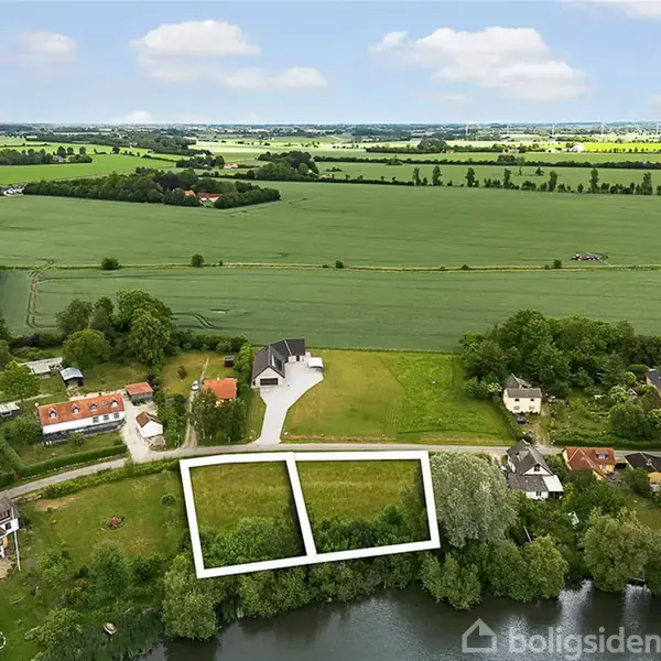 To grunde markeret med hvide linjer ligger tæt på en sø omgivet af landlige huse og åbne marker under en spredt, skyet himmel.