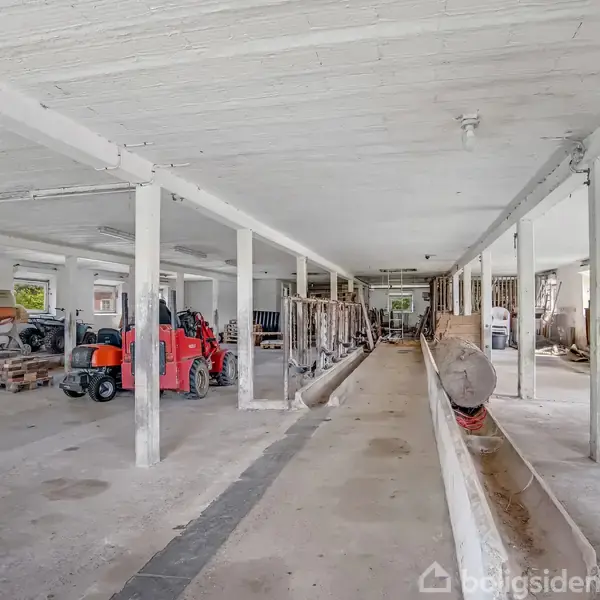 En tom lagerhal indeholder stillestående røde gaffeltrucks og byggematerialer. Kolonner deler rummet, der har en uafsluttet betonbund og store vinduer, der lukker naturligt lys ind.