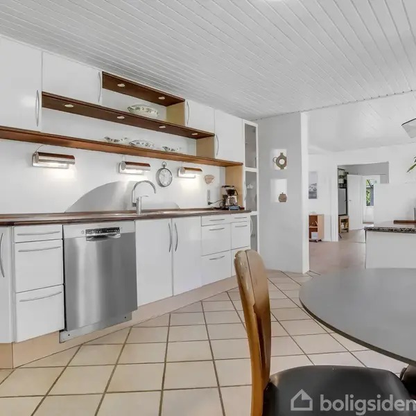 Køkken med hvide skabe og indbygget opvaskemaskine, bordplade med vask. Rummet er lyst og moderne med spisebord og stole i forgrunden, fliser på gulvet og et panoramavindue.