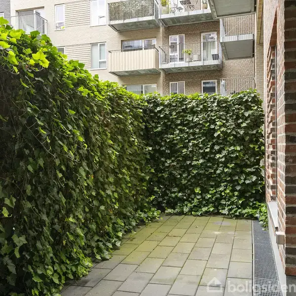 En lille, flisebelagt terrasse er omgivet af tæt, grøn beplantning. Til højre er der en murstensvæg, mens flere altaner fra en boligblok er synlige i baggrunden.