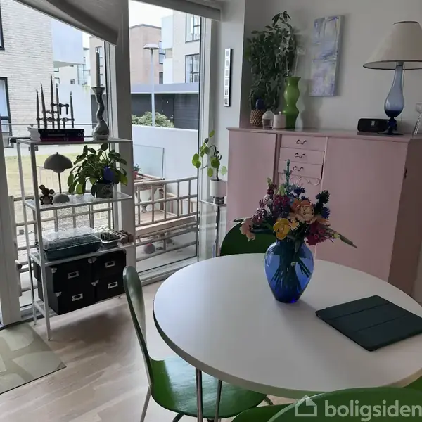 En rund hvid bord har en blå vase med blomster i et lyst rum. En reol med planter og figurer står ved en stor glasdør, der åbner til en terrasse.