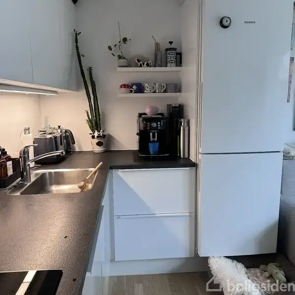 Hvidt køkken med sort bordplade. En kaffemaskine står ved siden af et køleskab. På hylder er der krus og planter. En vask med opvaskebørste er ved siden af en brødrister.