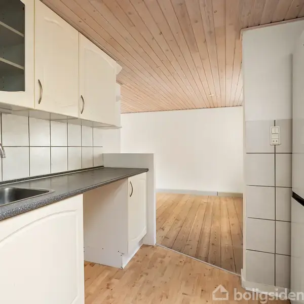 Et køkken med hvide skabe og en mørk bordplade, der strækker sig mod åbent stueområde med trægulve. Væggene er flisebelagte ved vasken.