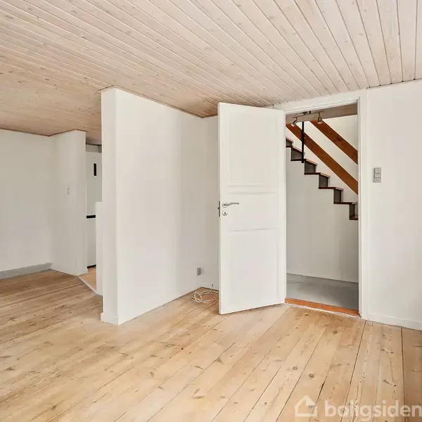 Et lyst rum med trægulv og træloft. En åben dør fører til en trappe i baggrunden. Vinduet til venstre har lette gardiner.