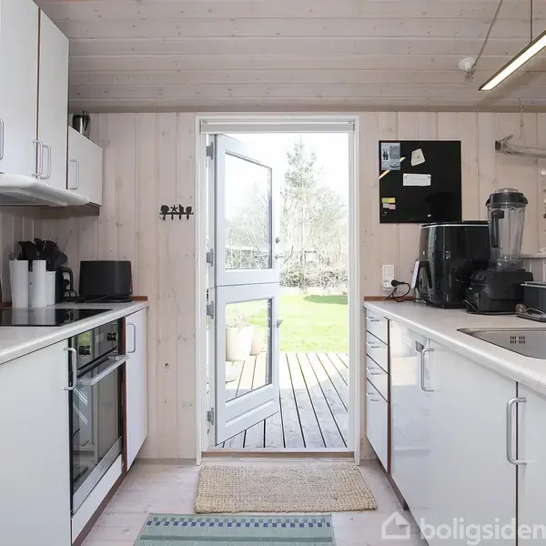 Køkken med hvide skabe og apparater, står åbent mod haven gennem en glasdør. En kaffemaskine og blender står på køkkenbordet. Rummelig og lyst miljø.