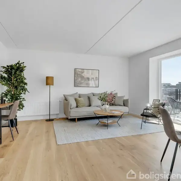 En stue med en grå sofa og sofabord på et tæppe, et skrivebord ved siden af en plante, samt spisebord med stole. Store vinduer giver udsigt til byen.