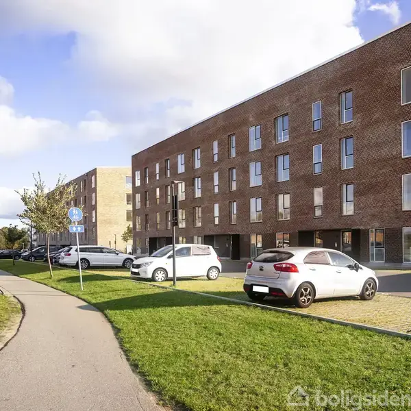 Murstensbygning står stille blandt parkerede biler og cykelsti. Græsplæne omgiver området og cykelparkering ses til venstre. Himlen er delvist overskyet.
