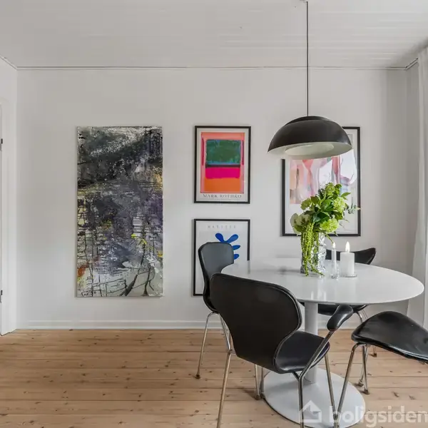 Rundt hvidt spisebord står i et lyst rum, omgivet af sorte stole. En stor lampe hænger over bordet. Væggen har kunstværker; et maleri, en plakat og abstrakt kunst.