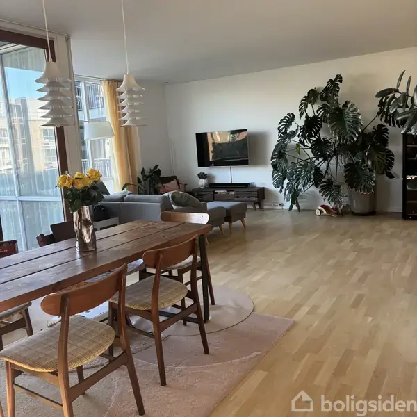 En stue med trægulv indeholder et træbord og stole med en vase med gule blomster. En sofa og et tv er placeret nær vinduerne. Store planter står i hjørnet.