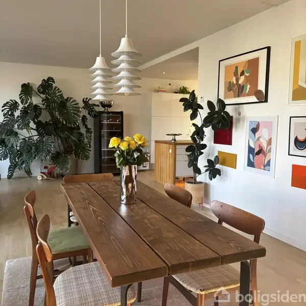 Et træbord med vase af gule blomster står i en lys spisestue, omkranset af stole og dekoreret med planter og farverig kunst på væggene.