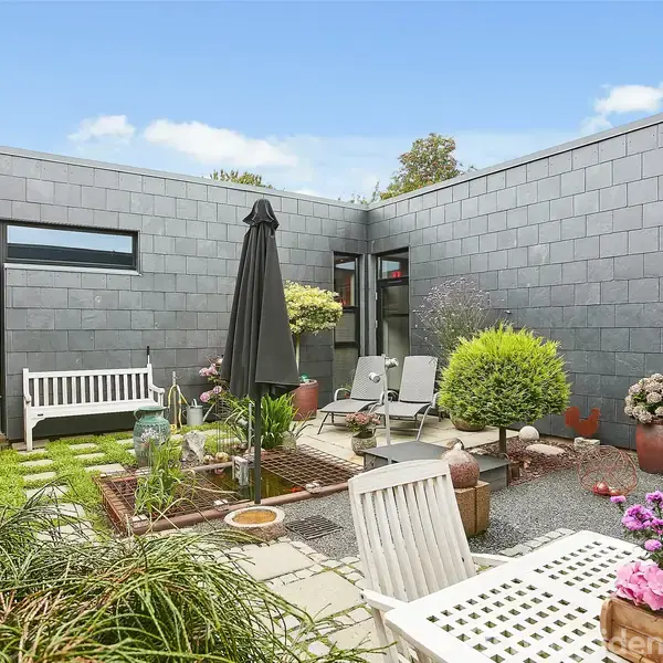 En gårdhave med hvidt havemøblement og planter, herunder en sort parasol. Baggrunden er en moderne grå murstensbygning med store vinduer. Blomster og grønne planter omkranser området.