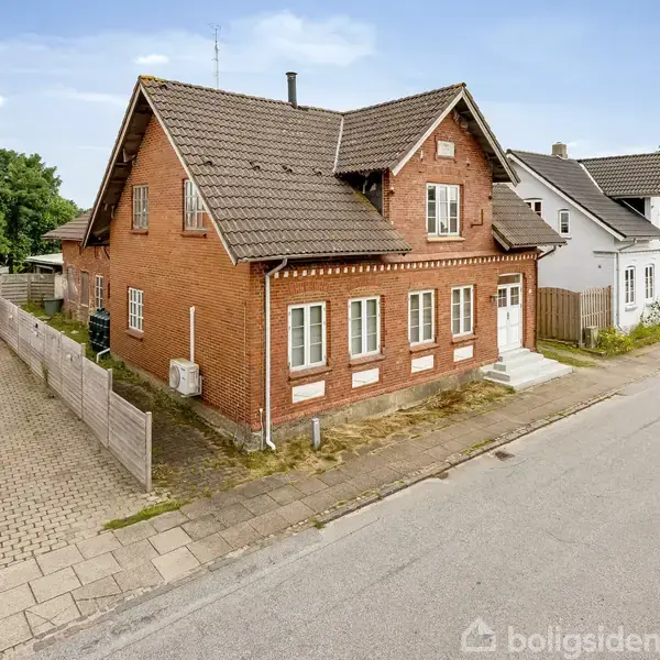 Rødt murstenshus med skråt tag og flere vinduer, står langs en stille vej mellem andre huse. En flisebelagt indkørsel fører til en bil ved siden af huset.