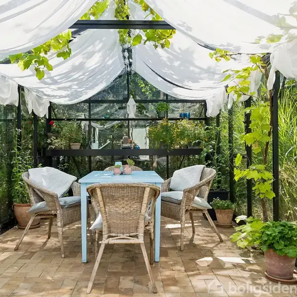 Moderne, sortmalet hus med fladt tag har en carport med en hvid bil parkeret. Indkørslen er omgivet af grønne planter og buske. Huset ligger i et beboelsesområde.