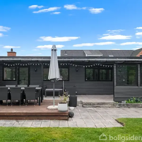 En mørkeblå stuehusvæg har store vinduer og en overdækket terrasse. På terrassen står et bord med sorte stole og en opstillet parasol. Græsplæne og fliser danner omgivelserne.