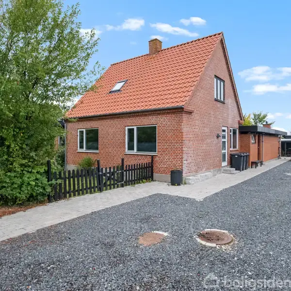 Et rødt murstenshus med rødt tegltag står på en grusbelagt indkørsel med omgivelser af træer og en hæk. En sort låge deler forhaven fra indkørslen.