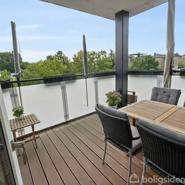 En altan med mørkt trægulv har et bord og stole, placeret til middagsselskab. Planter dekorerer rummet, som er delvist omgivet af glasværn. Omkring ses grønne trætoppe og bygninger.