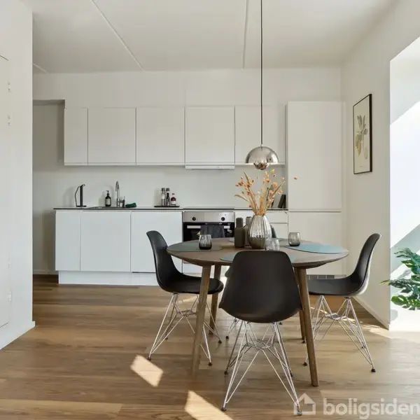 Et rundt spisebord med sorte stole står midt i et moderne køkkenalrum. En vase med tørrede blomster står på bordet, omgivet af hvide køkkenskabe og et stort vindue.