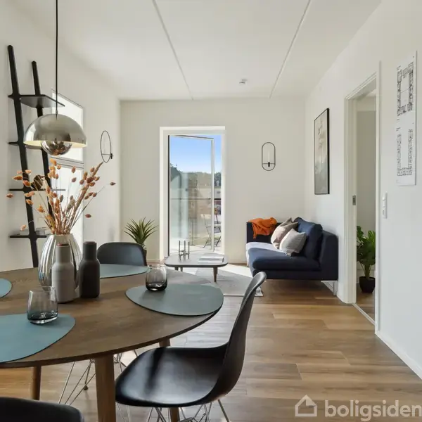 Rundt spisebord med dækkeservietter står i spisestue, omgivet af stole og dekorationer. I baggrunden ses en sort sofa og en lysfyldt altandør. På væggen hænger plantegninger.