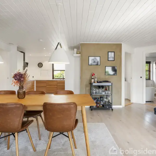Et spisebord med seks brune læderstole er placeret under to hvide pendellamper i en lys stue. Baggrunden har et barvogn, billeder på væggen, og et kig ind til andre rum.