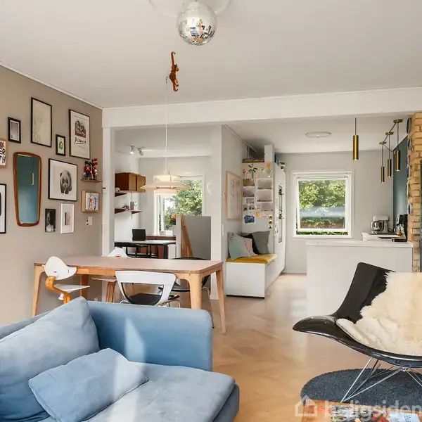 En stue med en blå sofa, betragtende et spisebord med stole. Væggen prydes af billeder i forskellige rammer. I baggrunden ses et lyst køkken med åbne skabe.