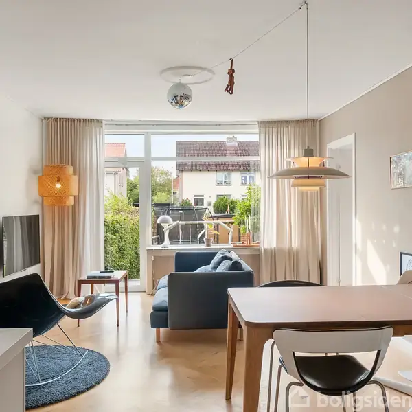 Stue med blå lænestol, der vender mod en fjernsynsskærm på væggen. Bordlampe og pendellampe lyser rummet. Store vinduer med udsigt til en have bagved. Vægdekorationer hænger rundt omkring.