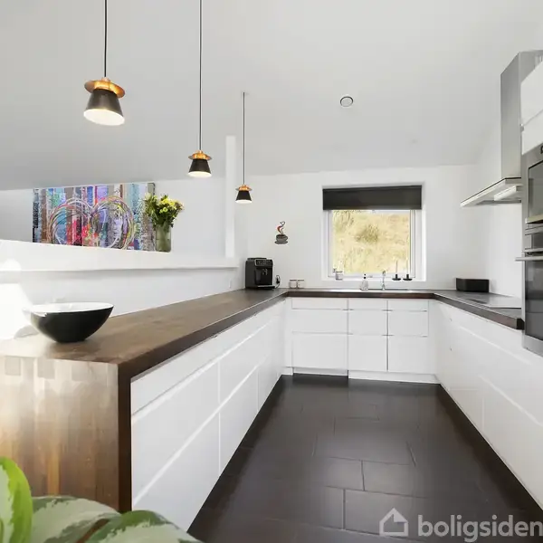 Moderne køkken med hvide skabe og mørkebrun bordplade, tre hængende lamper. Køkkenmaskiner indbygget. Et vindue med sort rullegardin og planterummer tilføjer farve.