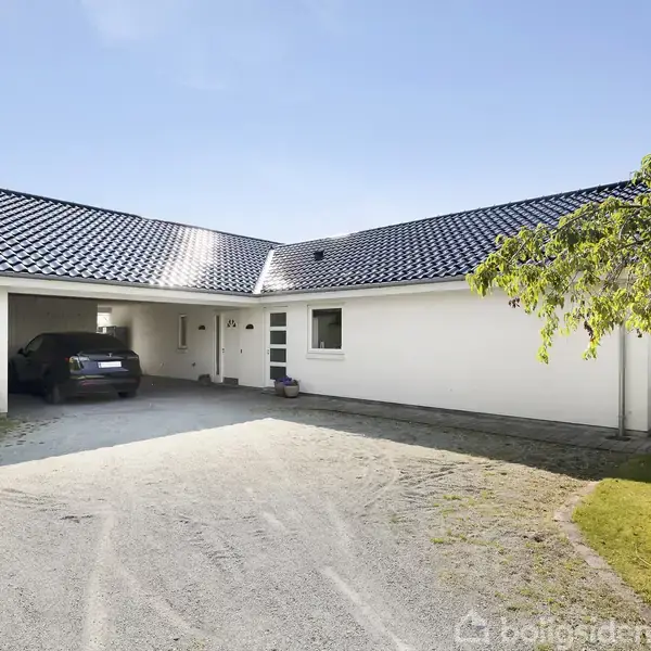 Hvid bungalow med sort tegltag. En bil holder parkeret i carporten. Omgivet af en have med græs og et træ til højre. Klar himmel i baggrunden.