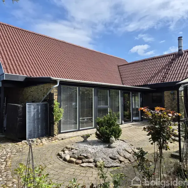 Hus med rødt tegltag og store glaspartier, omkranset af flisebelagt gårdhave med planter og blomsterbed under en blå himmel. En enkelt træbænk står ved siden af bygningen.
