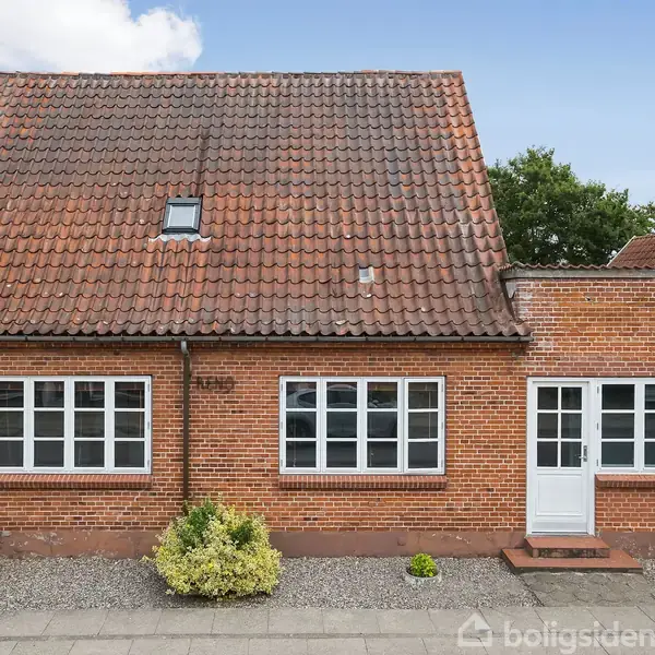 Et rødt murstenshus med et skråt, teglbelagt tag har to store vinduer og en hvid dør foran. Foran huset er der en grusbelagt have med nogle buske.