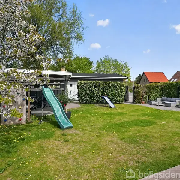 Et grønt legeområde med en rutsjebane i en have med græsplæne, omgivet af blomstrende træer og hække. En terrasse med havemøbler ses i baggrunden under en blå himmel.