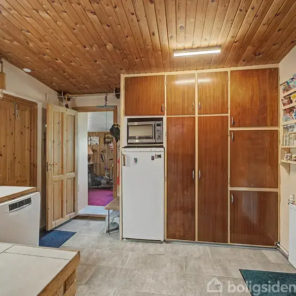 Køkken med træloft og store skabe i træ, indeholder mikroovn og køleskab. Vægge har hylder med souvenirs. Døråbning fører til anden del af huset. Møbler og radiator ses.