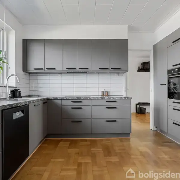 Gråt køkken med L-formet bordplade. Skabslågerne er mørkegrå, og hvide fliser pryder væggen. En grøn plante står ved vinduet, og der er et indbygget komfur.