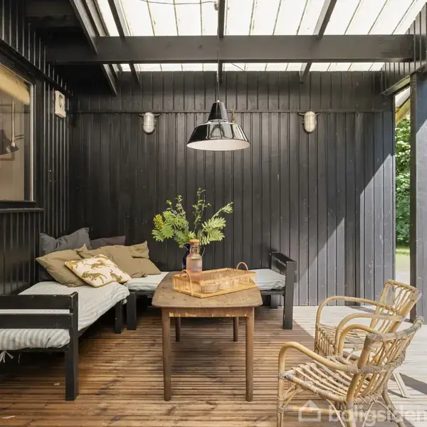 Indendørs siddeområde med sorte trævægge og træmøbler. To sofaer med puder står rundt om et træbord med en plante og bakke. Nuancer af sort skaber en hyggelig atmosfære.