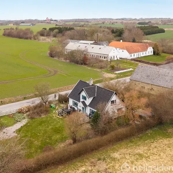 Et hvidt hus med sort tag og veranda står omgivet af buske og træer. En vej løber forbi, mens bygninger og marker strækker sig i baggrunden under en lys himmel.
