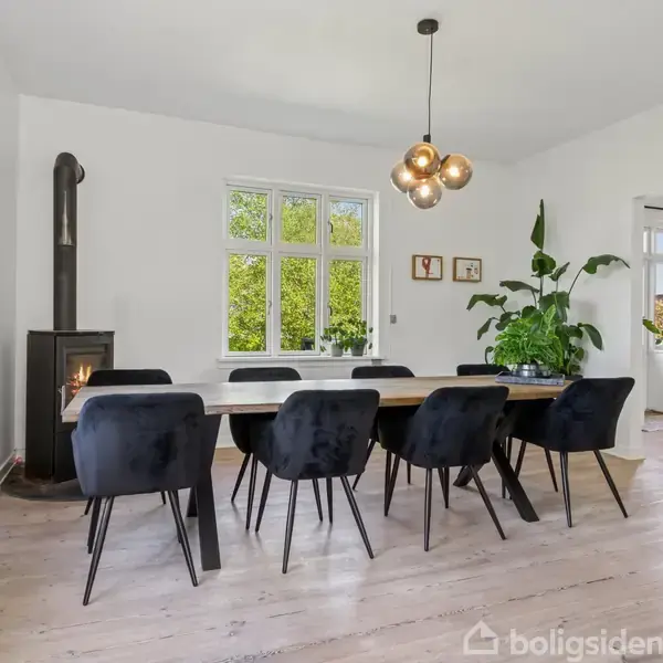 En spisebord med otte sorte stole står i en lys spisestue. Langs væggen ses en brændeovn og store grønne planter. Vinduerne viser udsigt til grønne træer.