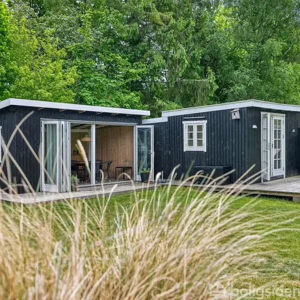 Sort træhus med åbne glasdøre står på en træterrasse, omgivet af en grøn græsplæne og frodig skov. En liggestol er placeret på terrassen til højre.
