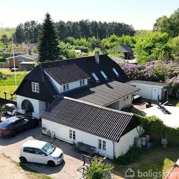 Et stort hus med sort tag står i et grønt område med træer og buske. Biler er parkeret på grusvejen ved siden af huset, hvor solen skinner.