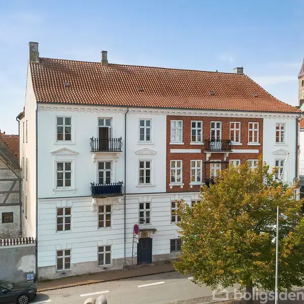Et stort, hvidt og rødt murstenshus med altaner ses fra gadeniveau. Træer og biler omkranser bygningen, mens en kirketårn er synlig i baggrunden under en blå himmel.